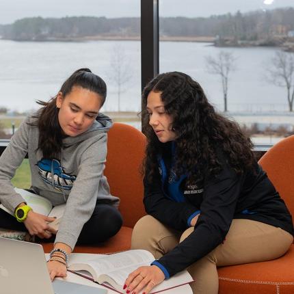 Two students studying in front of a large window in the Commons that overlooks the Saco River