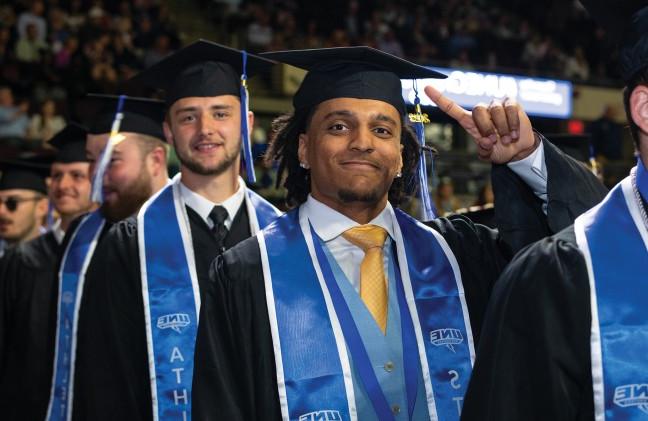 A student in academic regalia points to his black cap at graduation